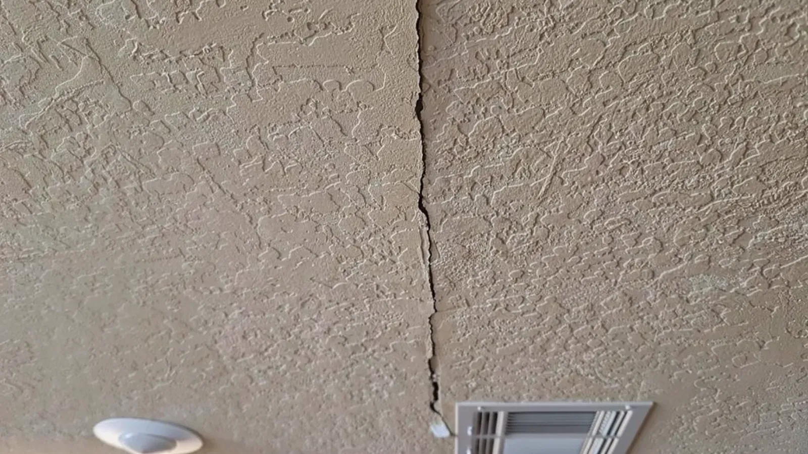 A crack split open along a joint in a textured painted ceiling, running past a round recessed light and a rectangular ceiling air register