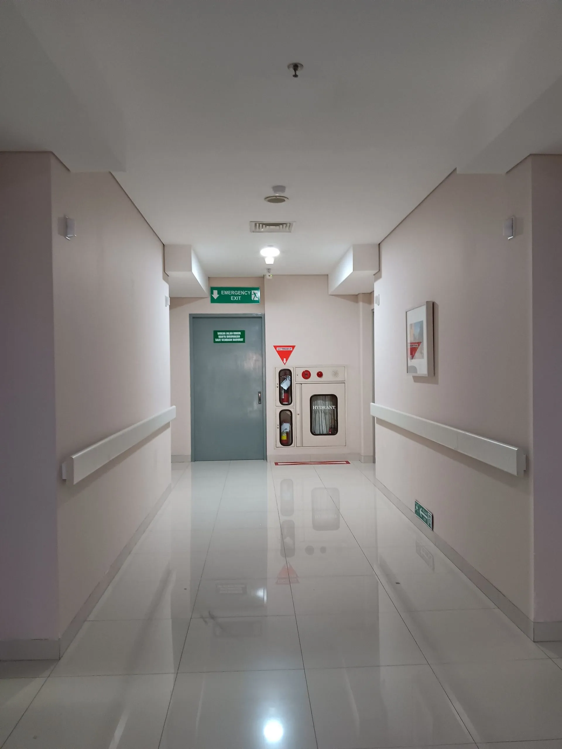 Finished medical facility corridor with smooth painted drywall walls, wall-mounted handrails, recessed lighting, a glossy white tile floor, and an illuminated emergency exit sign