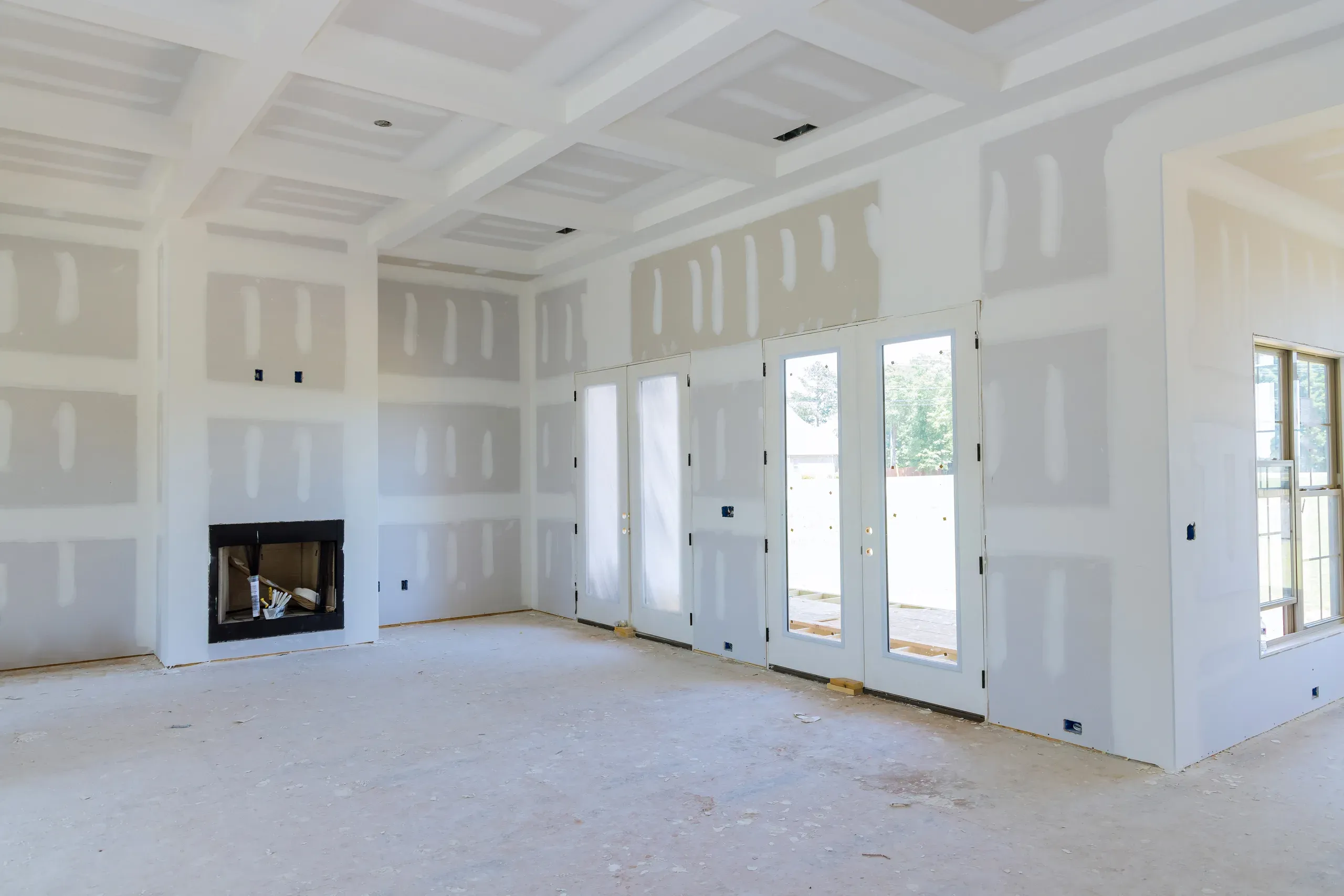 Large great room with a coffered drywall ceiling, seams taped and coated and fasteners spotted throughout, a bare firebox opening and French doors with sidelights, dust on the unfinished floor