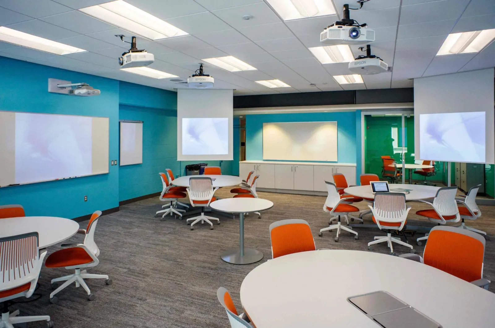 Modern collaborative classroom with finished painted drywall walls in teal, ceiling-mounted projectors, wall-mounted whiteboards and projection screens, and clustered round tables with orange task chairs