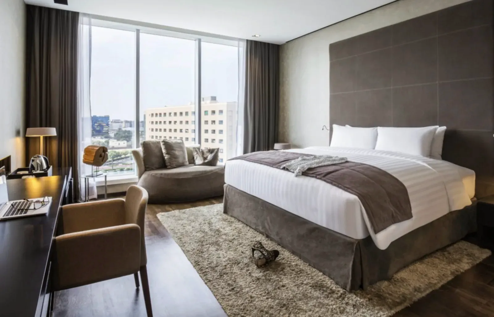 Finished hotel guest room with smooth painted drywall walls and ceiling, an upholstered headboard, a sheer-curtained window, and a padded bench at the foot of the bed