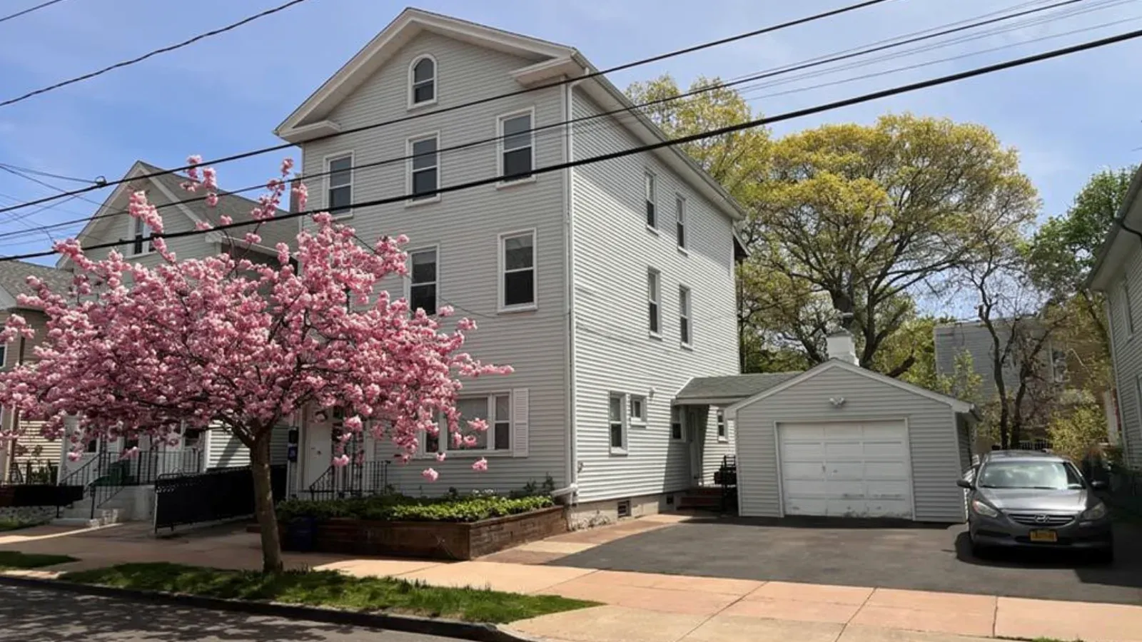 A three-story multi-family house with grey clapboard siding, a detached garage and a flowering tree out front