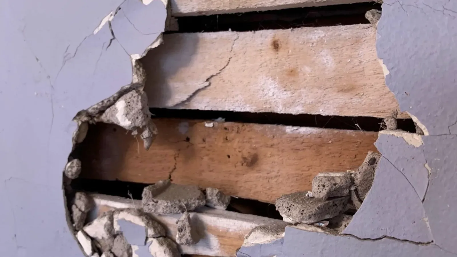 Hole broken through a painted plaster wall, exposing the horizontal wood lath behind it and the crumbling plaster keys around the opening