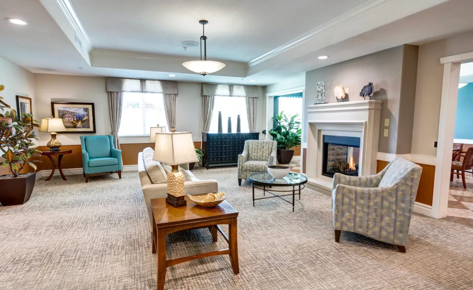 Senior-living community common lounge with finished painted drywall, crown molding, a tray ceiling, a stone fireplace, and upholstered armchairs arranged around a coffee table